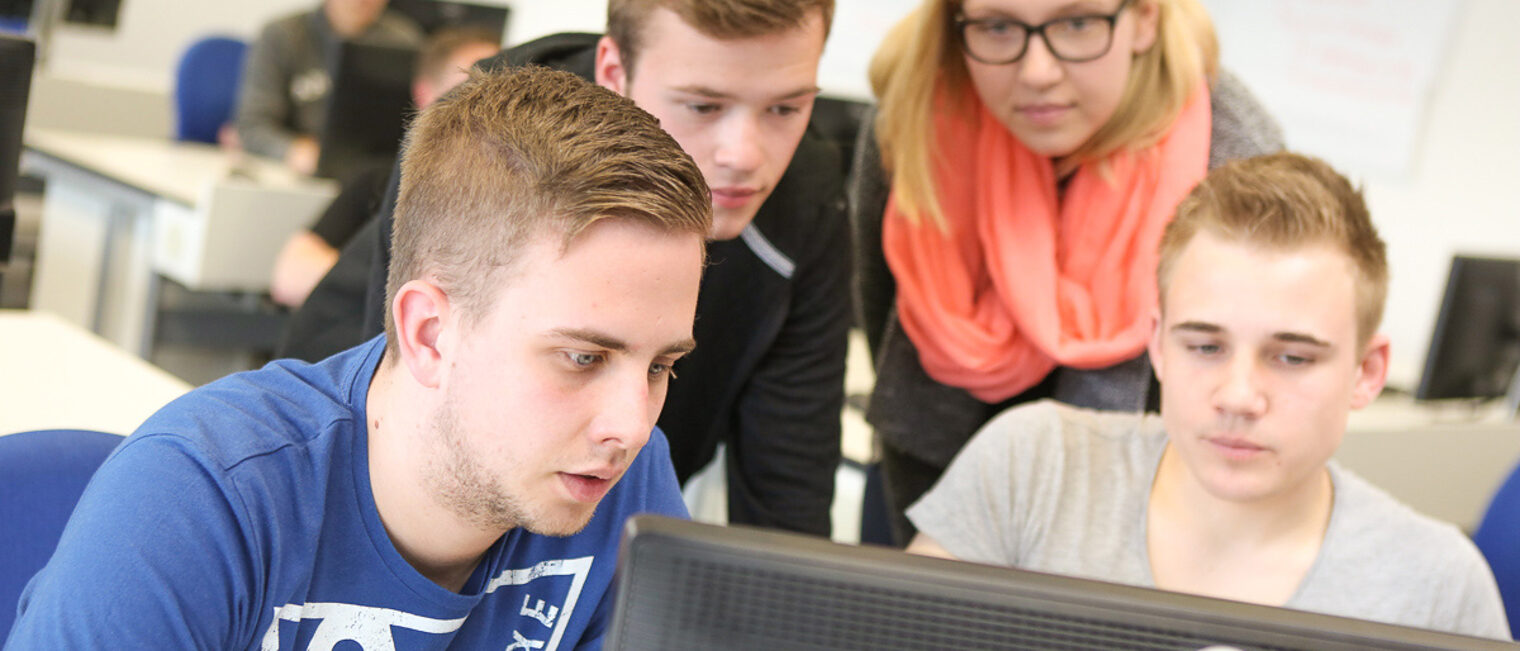 Kursteilnehmer sitzen vor einem Computer. Eine Kursteilnehmerin blickt ihnen dabei über die Schulter.