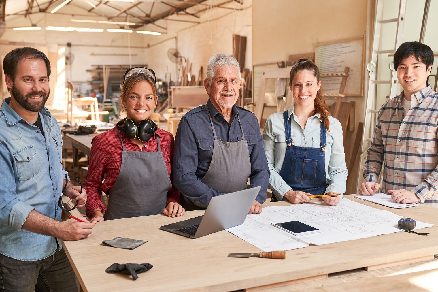 Gruppe von Tischlern stehen an einem Tisch in einer Werkstatt und lächeln in die Kamera.