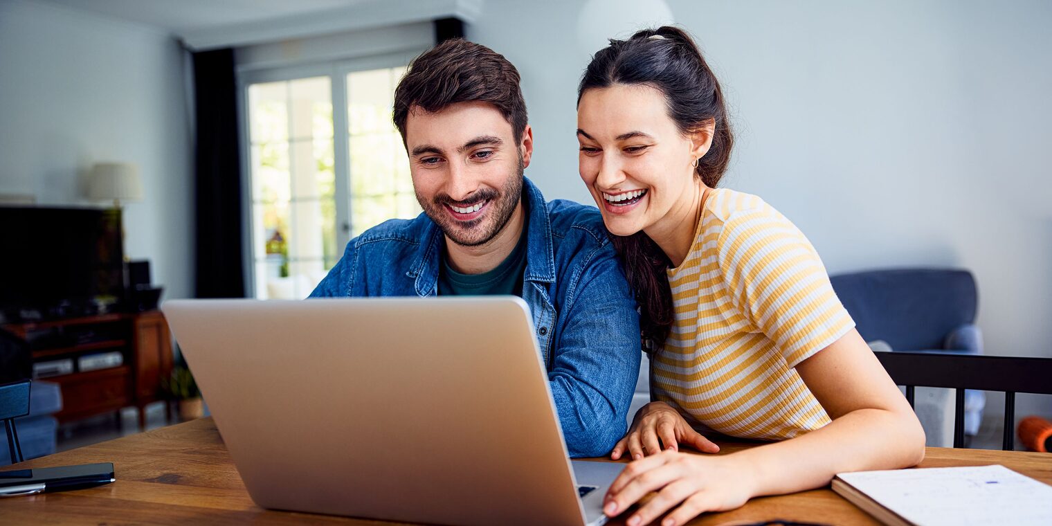 Junge Frau und junger Mann sitzen zusammen an einem Tisch und blicken mit Freude in einen Laptop.