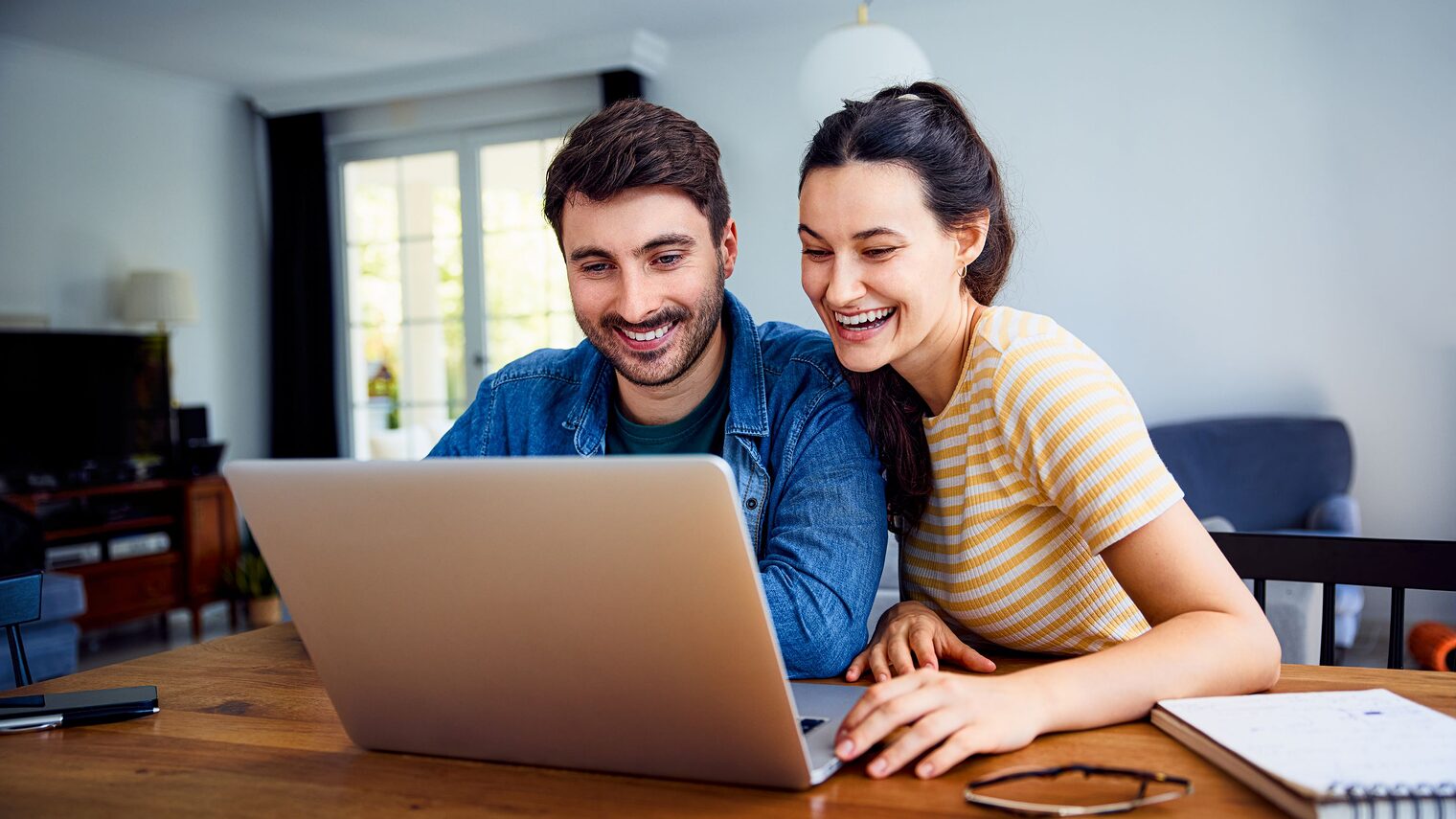 Junge Frau und junger Mann sitzen zusammen an einem Tisch und blicken mit Freude in einen Laptop.