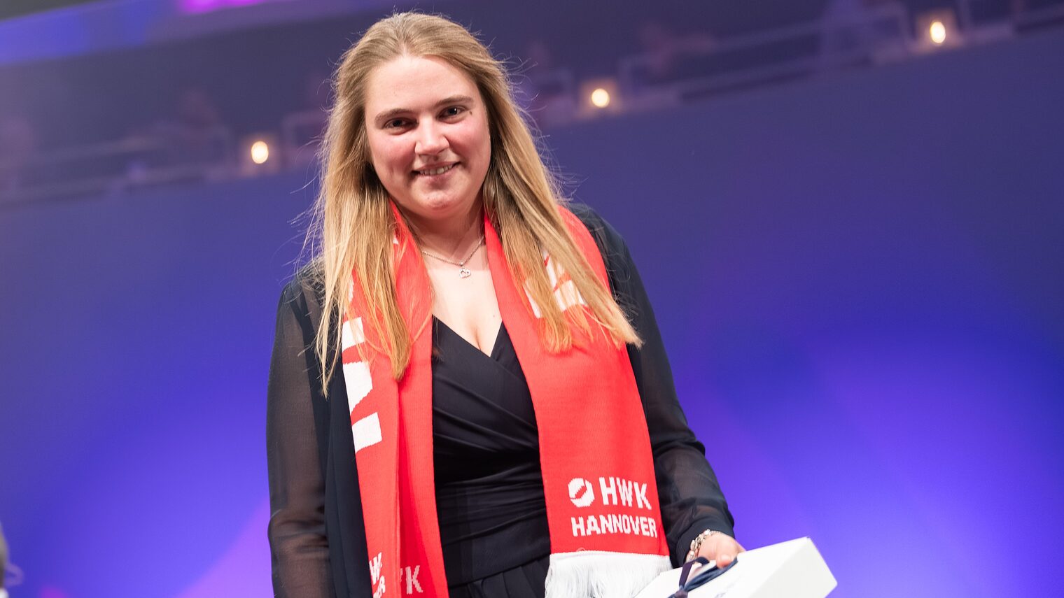 Porträt von Schornsteinfegermeisterin Rieke Bößling bei der Meisterfeier 2025 mit Meister-Schal und Geschenk im Scheinwerferlicht.