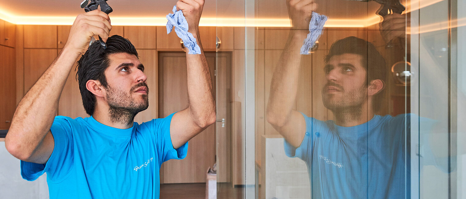 Ein Handwerker trägt sorgfältig Dichtmasse an einer Glaswand auf. Mann im blauen T-Shirt benutzt eine Kartuschenpistole, um Dichtmasse an einer Glaswand aufzutragen; seine Arbeit spiegelt sich im Glas wider.