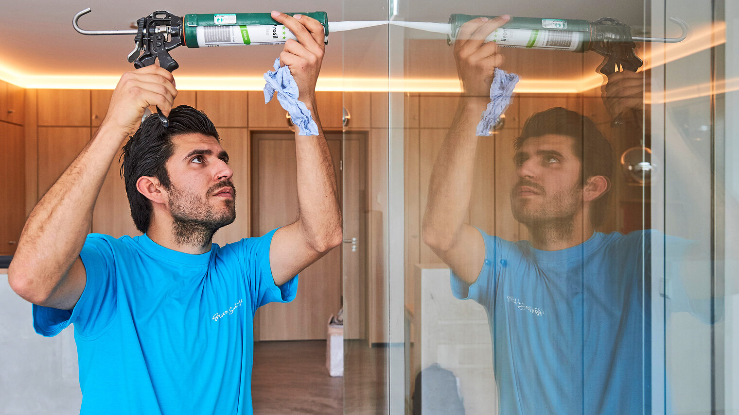 Mann im blauen T-Shirt benutzt eine Kartuschenpistole, um Dichtmasse an einer Glaswand aufzutragen; seine Arbeit spiegelt sich im Glas wider.