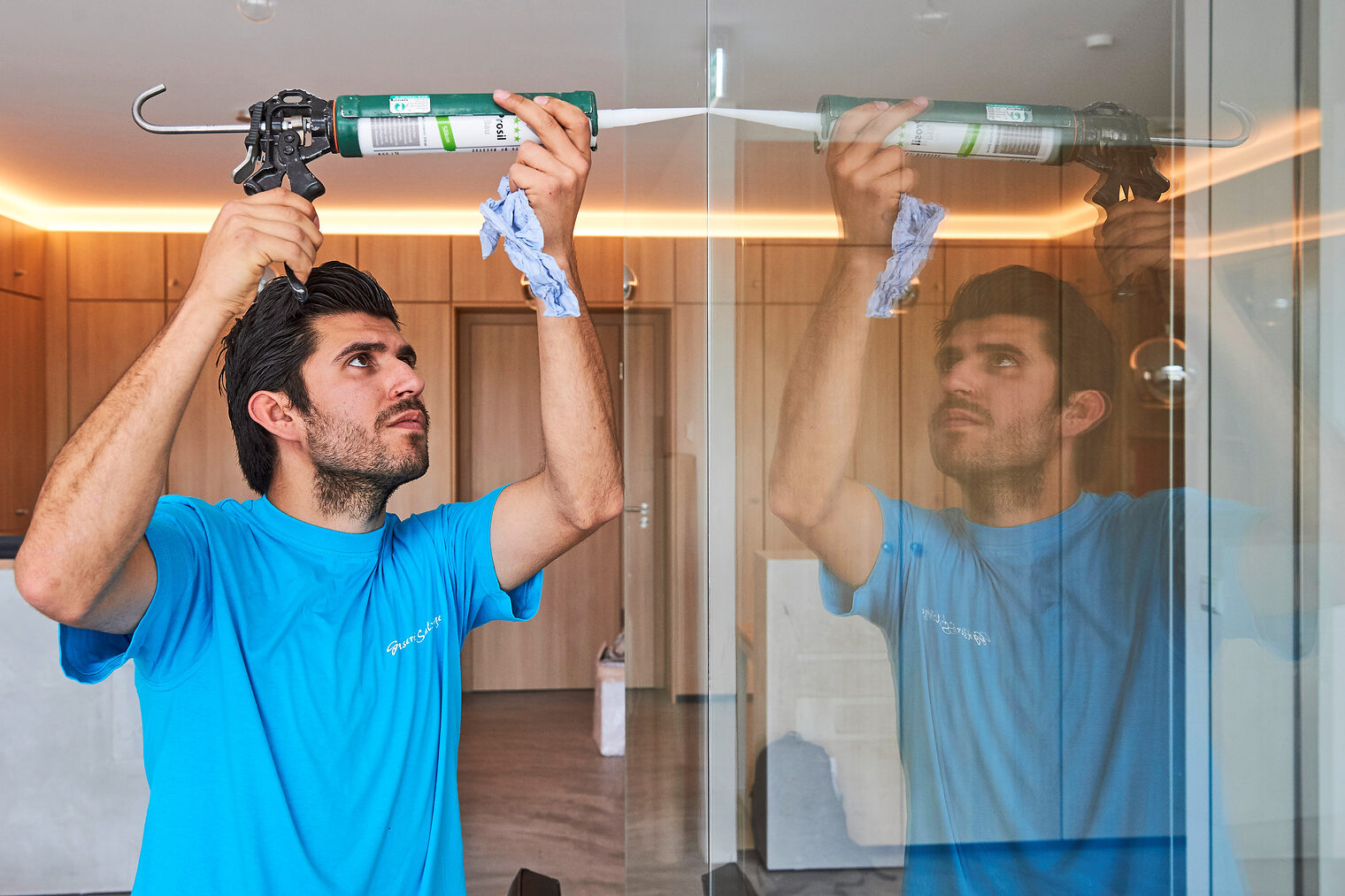 Mann im blauen T-Shirt benutzt eine Kartuschenpistole, um Dichtmasse an einer Glaswand aufzutragen; seine Arbeit spiegelt sich im Glas wider.