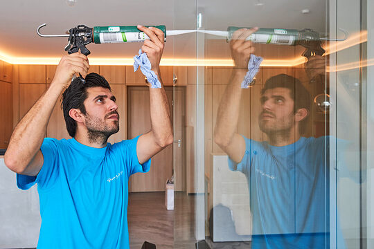 Mann im blauen T-Shirt benutzt eine Kartuschenpistole, um Dichtmasse an einer Glaswand aufzutragen; seine Arbeit spiegelt sich im Glas wider.