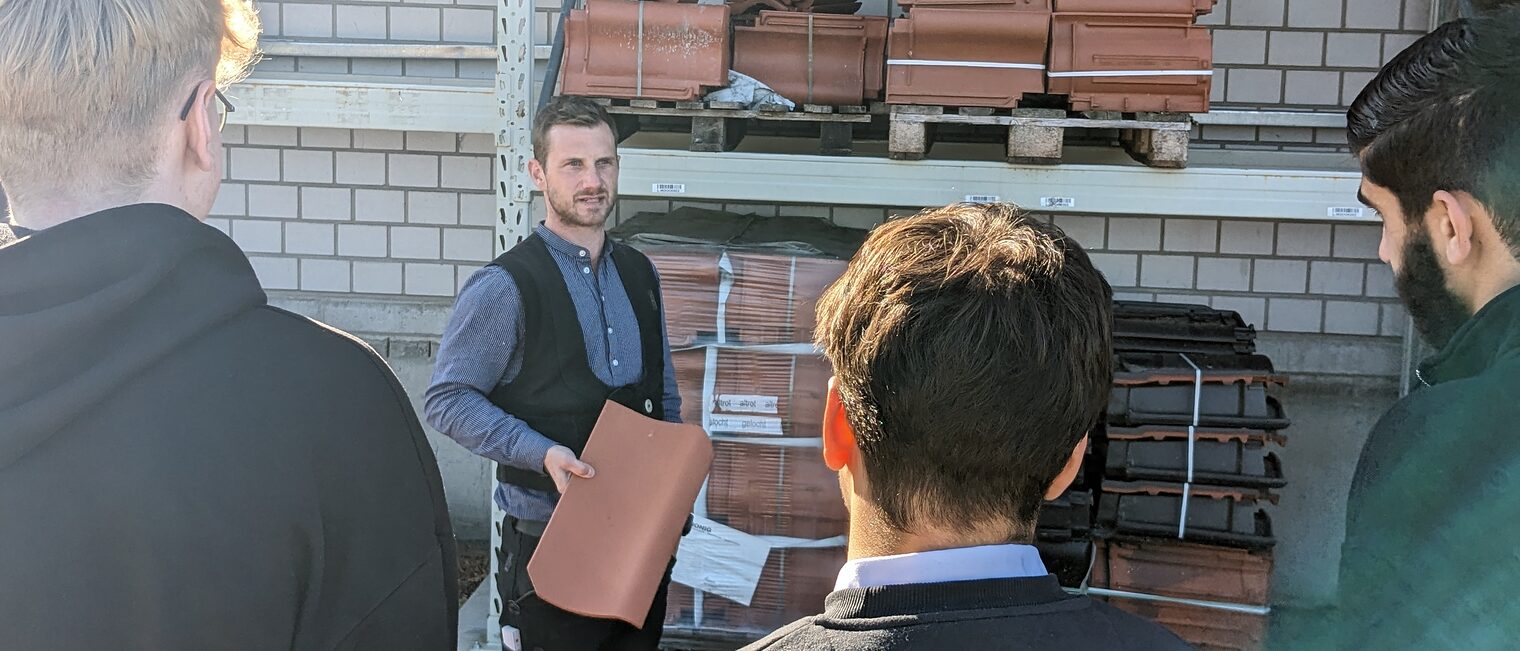 Dachdeckermeister Florian Kastner zeigt einen Ziegel, Jugendliche schauen zu.