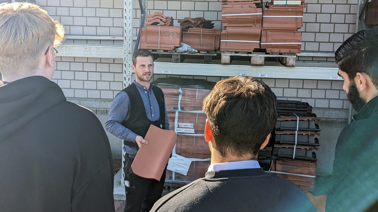 Dachdeckermeister Florian Kastner zeigt einen Ziegel, Jugendliche schauen zu.