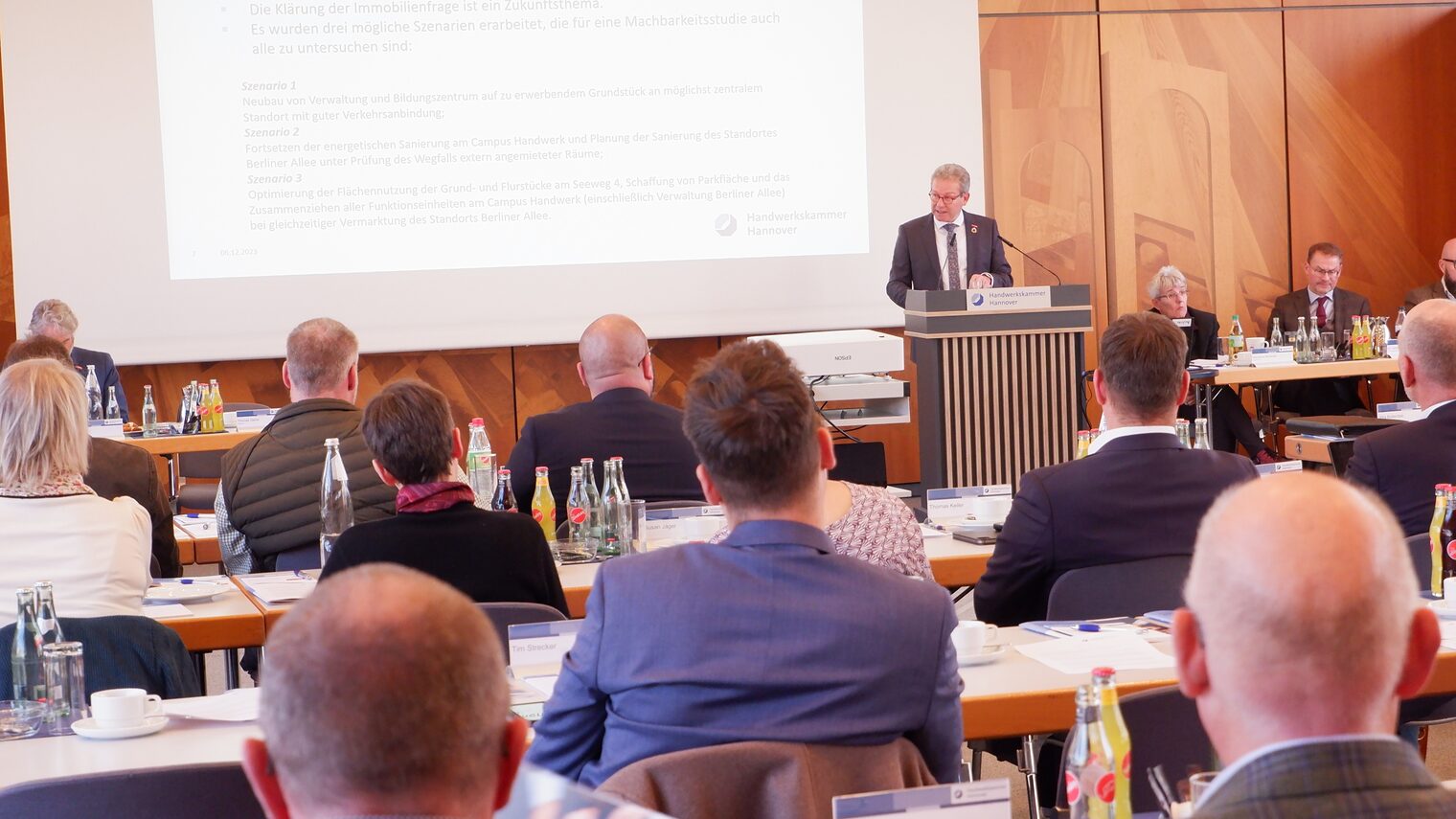 Mann am Pult spricht vor Menschen an Tischen.