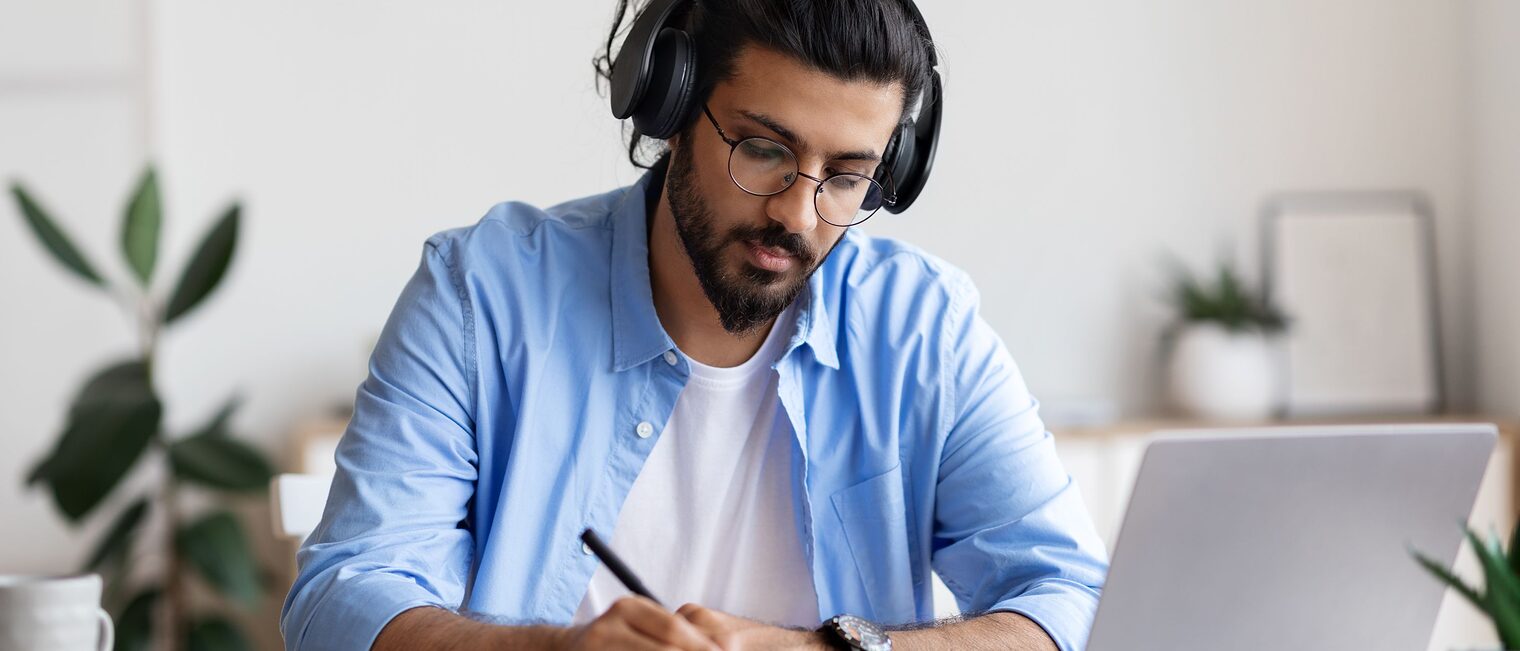 Junger Mann sitzt mit einem Laptop und Kopfhörer am Tisch und notiert sich etwas auf Papier.