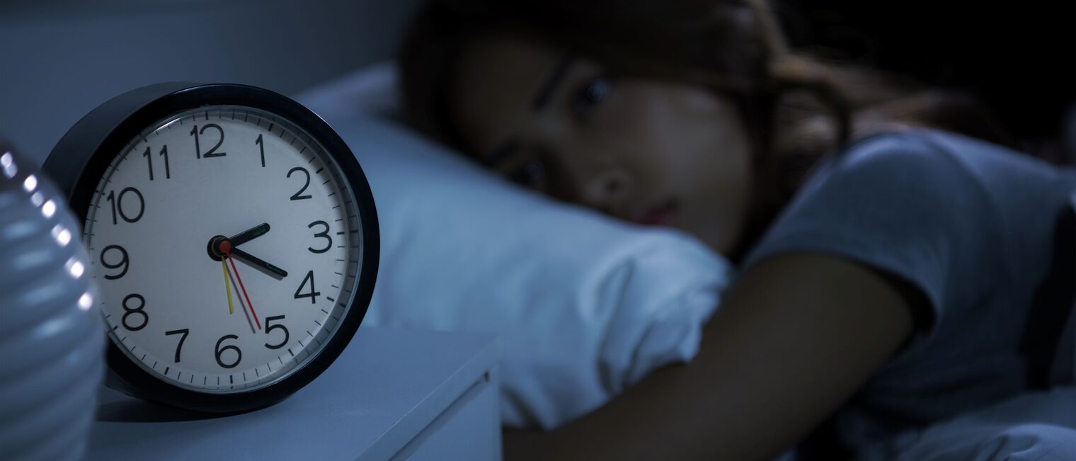 Frau liegt nachts wach im Bett, neben ihr auf dem Nachttisch ein Wecker, der die Uhrzeit 02:20 Uhr anzeigt. 