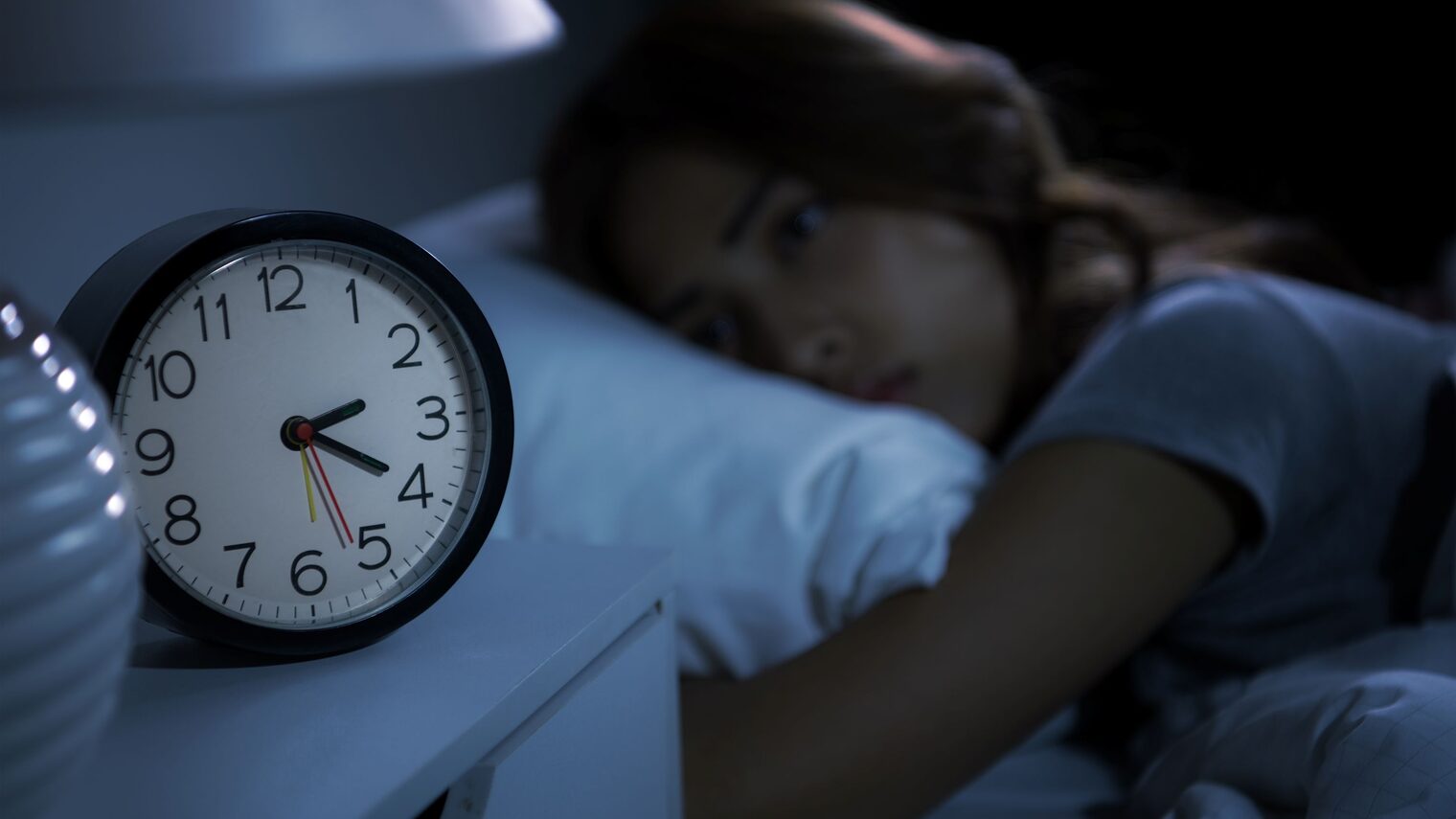 Frau liegt nachts wach im Bett, neben ihr auf dem Nachttisch ein Wecker, der die Uhrzeit 02:20 Uhr anzeigt. 