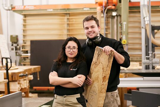 junge Frau und junger Mann nebeneinander in einer Tischlerwerkstatt.