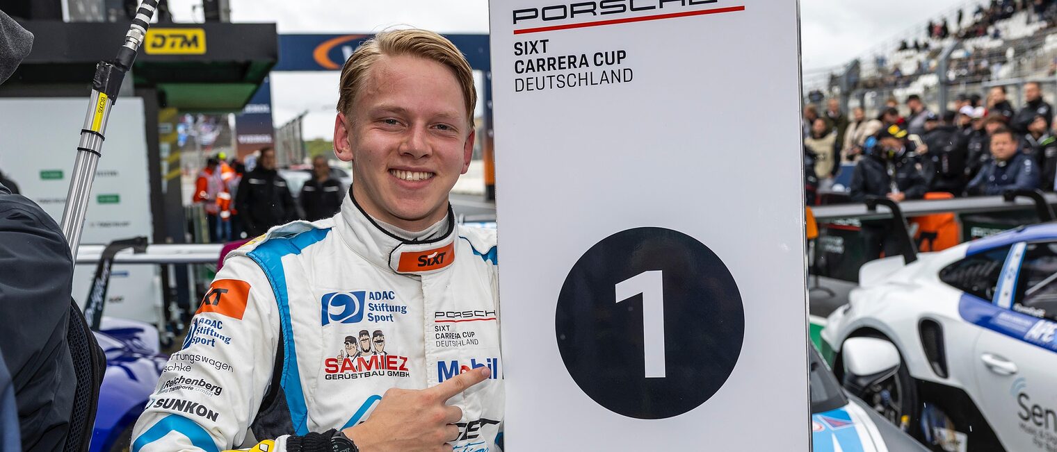 Janne Stiak am Rande der Rennstrecke zeigt auf ein Banner mit dem Aufdruck Nummer 1 beim PorscheCup.