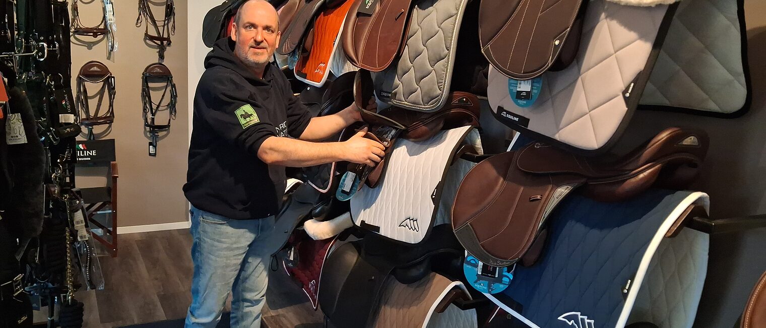 Jens Jadischke mit Blick in die Kamera an einer Reihe von Reitsportsätteln.