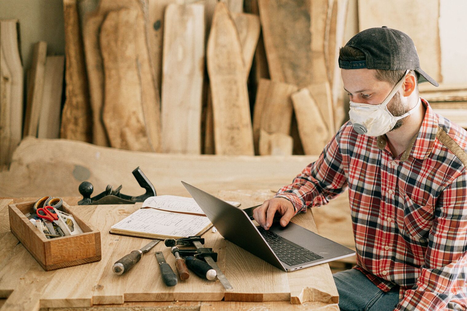 Handwerker sitzt mit FFP-Maske an seinem Laptop in der Werkstatt. Auf dem Tisch liegen Werkzeuge und ein Notizbuch.