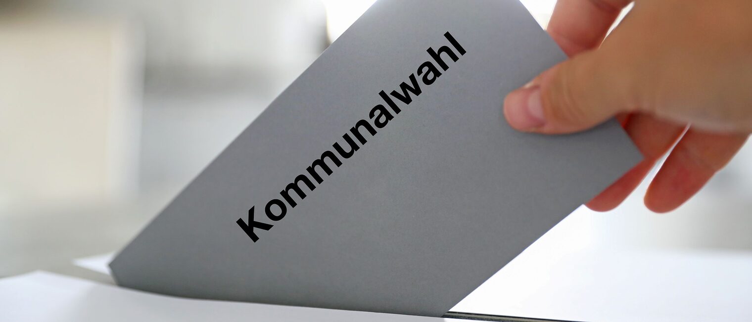 Hand steckt Wahlzettel mit Kommunalwahl-Aufschrift in Wahlurne.