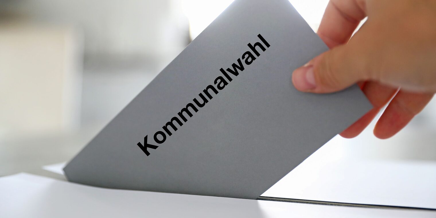 Hand steckt Wahlzettel mit Kommunalwahl-Aufschrift in Wahlurne.