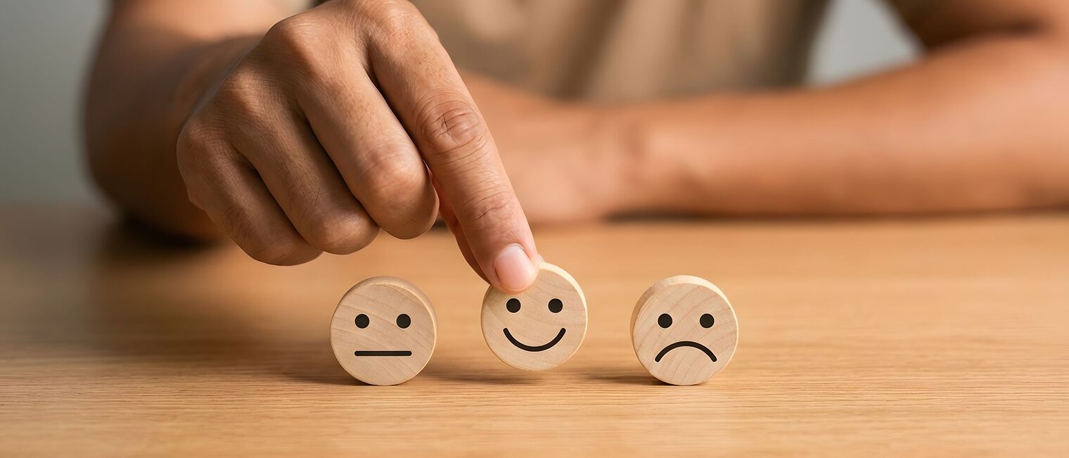 Drei Holzsmileys mit unterschiedlichen Gesichtsausdrücken werden von einer Hand gehalten.