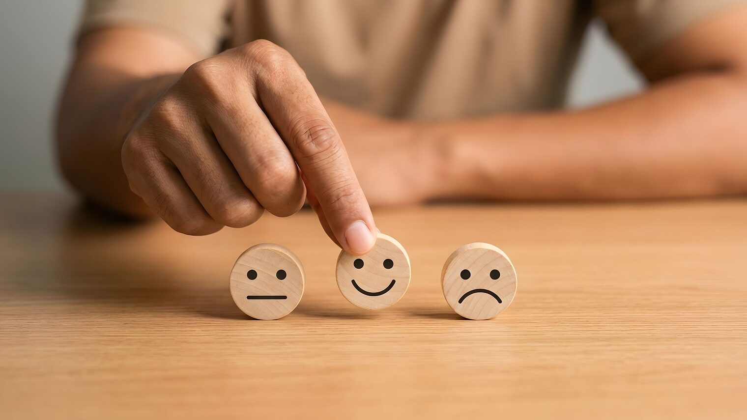 Drei Holzsmileys mit unterschiedlichen Gesichtsausdrücken werden von einer Hand gehalten.