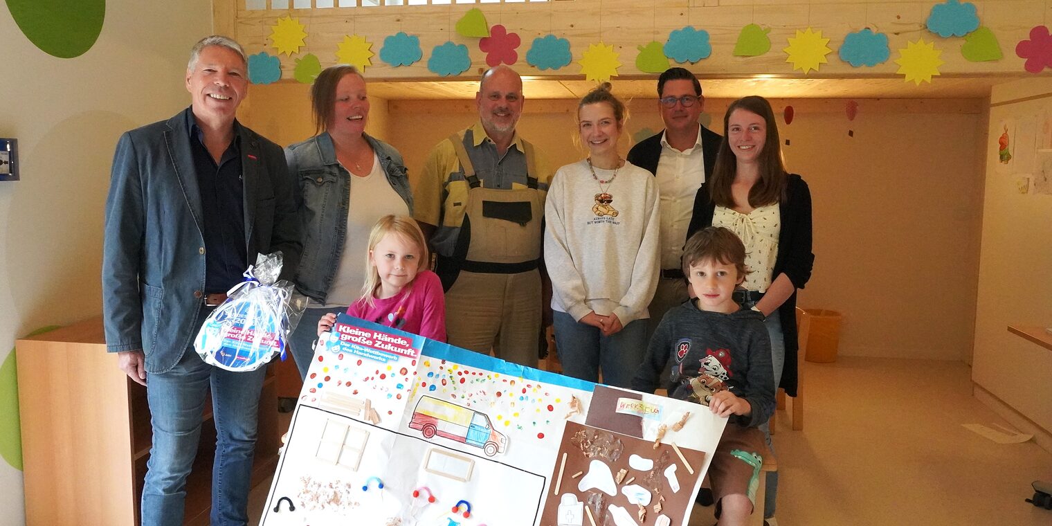 Fünf Erwachsene und zwei Kita-Kinder präsentieren das Gewinner-Plakat und die Plakette.