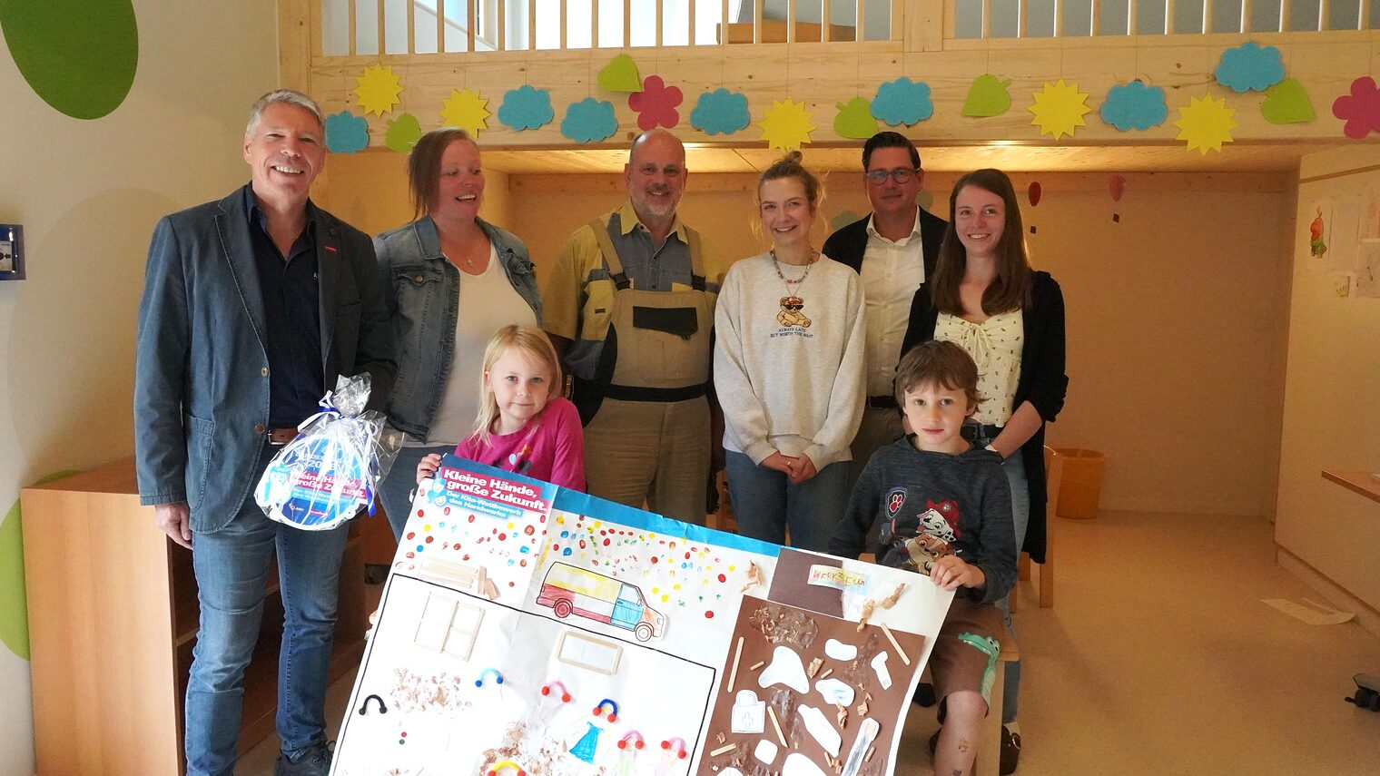 Fünf Erwachsene und zwei Kita-Kinder präsentieren das Gewinner-Plakat und die Plakette.