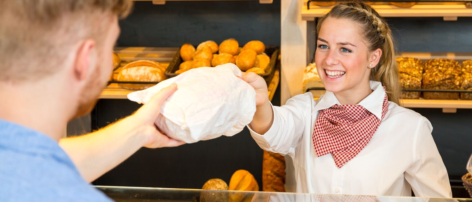 Eine Bäckereifachverkäuferin reicht eine gefüllte Brötchentüte über den Tresen.
